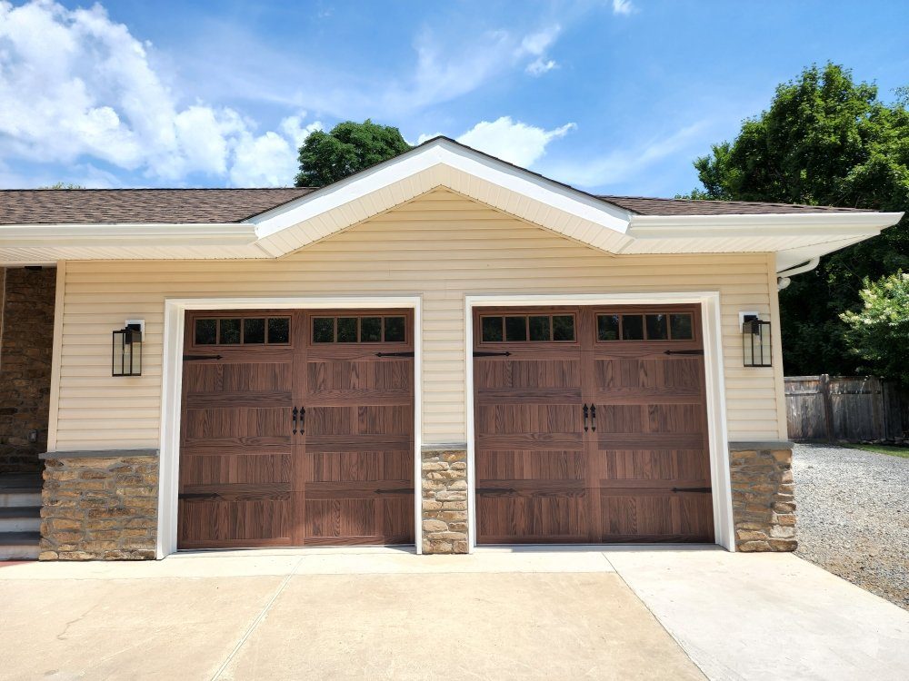 Two walnut wood-grain steel carriage doors with decorative hardware on a stone-front home